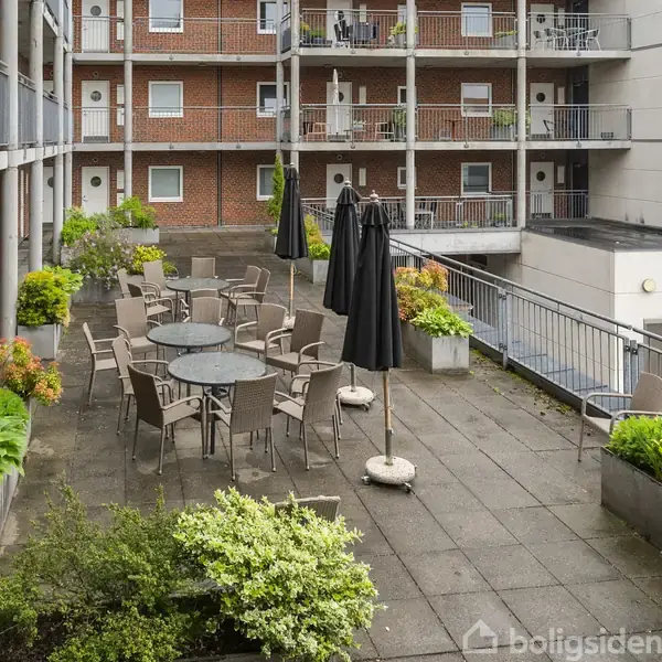 Udendørs terrasse med runde borde og stole står stille omgivet af grønne plantekasser. I baggrunden ses en murstensbygning med altaner.