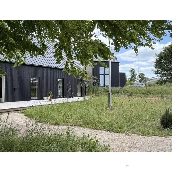 Sort hus med skrå tag står stille i en frodig have med træer og græs. En grussti snor sig gennem haven. Himlen er delvist overskyet.