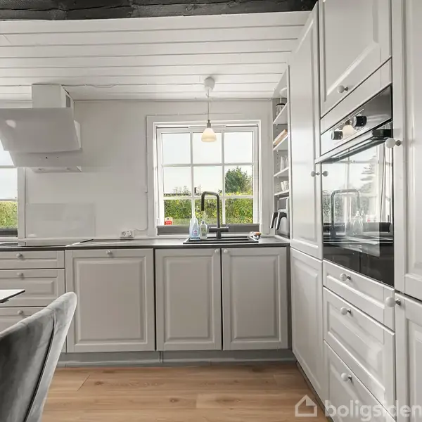 Et moderne køkken med hvide skabe, en ovn, og et stort vindue. En køkkenvask er i midten, omgivet af natur udenfor. Træbordet har grå stole ved siden af.