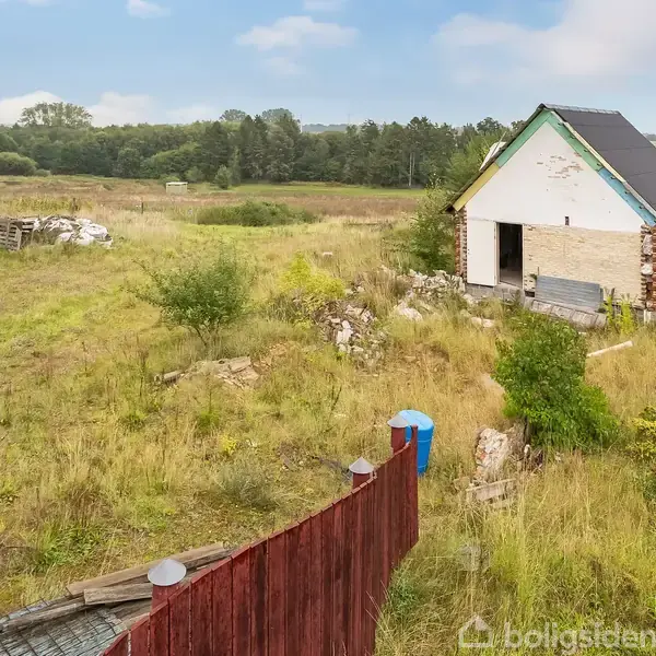 Et lille, ufuldstændigt hus står ensomt på en overgroet grund med højt græs og spredte buske. I baggrunden ses en skov, og der er en rød hegnssektion foran.