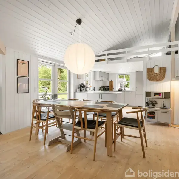 Et lyst, stilrent køkken-alrum med spisebord og stole i træ. En stor, rund lampe hænger over bordet. Rummet har en åben hems og en trappe samt store vinduer mod en grøn have.