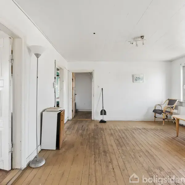 Stue med trægulv og hvide vægge, indeholdende en åben dør, en gulvlampe, en reol, og to vinduer. En stol og et bord står ved vinduet. Kontext af et enkelt og lyst rum.
