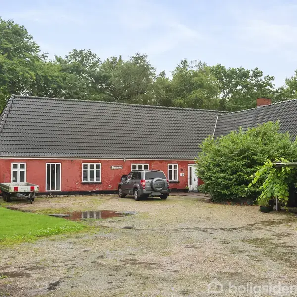 Rødt murstenshus med mørkt tag står i en landlig have. En bil holder parkeret foran, og der er en grusvej. Træer omkranser stedet, og et skur ses til højre.