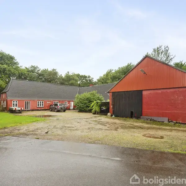 Rød ladebygning med store døre står på en gårdsplads omgivet af grønne træer og buske. En firkantet gårdbygning ses til venstre under en overskyet himmel.