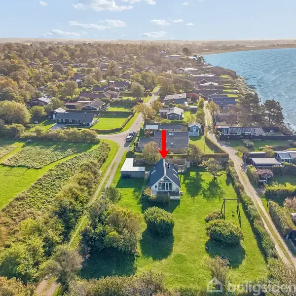 Hus markeret med rød pil, står i en grøn have tæt på en kystlinje. Omkringliggende område består af flere huse og rigeligt grønt landskab ved siden af havet.