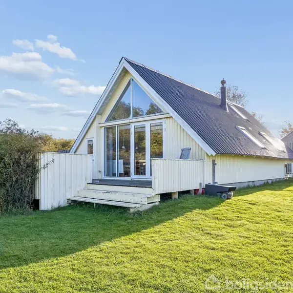 Et moderne, trekantet sommerhus med store glasdøre ligger på en velplejet græsplæne. Omkranset af buske og træer, er huset badet i sollys under en klar blå himmel.