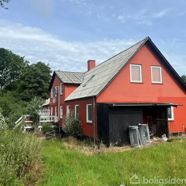 Rødt hus med skråtag står stille i en frodig have omkranset af tæt grønt løv og træer, mens skraldespande ses ved en overdækket bygning ved siden af huset.