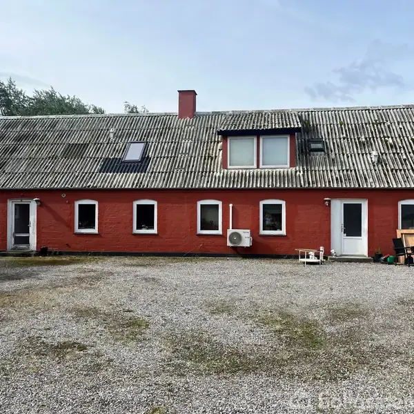 Rødt hus med skråt tag, flere vinduer og døre. Grå grusplads foran, omgivet af træer. En luftkonditioneringsenhed ved væggen.