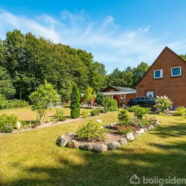 Rødt murstenshus står stille i en solrig have med velplejede blomsterbede og græsplæne. En mørk bil er parkeret ved siden af huset, omgivet af grøn skov i baggrunden.