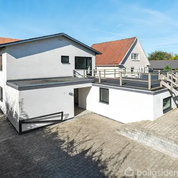 En hvid, moderne to-etagers villa står stille, omgivet af en brostensbelagt indkørsel og en trappe til en terrasse. I baggrunden ses andre huse og en blå himmel.