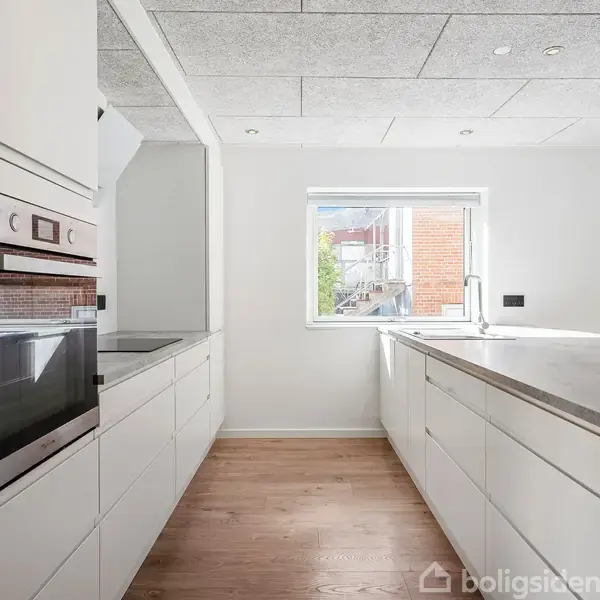 Et moderne køkken med hvide skabe og skuffer omgiver en indbygget ovn til venstre. Et stort vindue lyser rummet op, og en køkkenø står centralt på trægulvet.