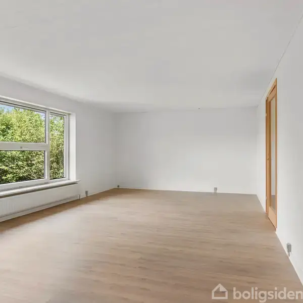 Et lyst og tomt værelse med trægulv, hvide vægge, og et stort vindue til venstre, der viser grønne træer udenfor. En trædør ses til højre.