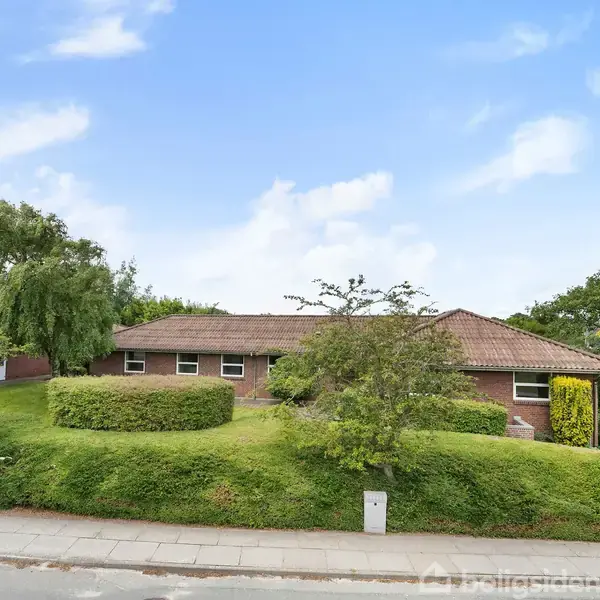 Hus af mursten med rødt tag ligger på en grøn bakke omgivet af buske og træer. En grå fortov langs vejen i forgrunden. Skyer spredt på blå himmel.