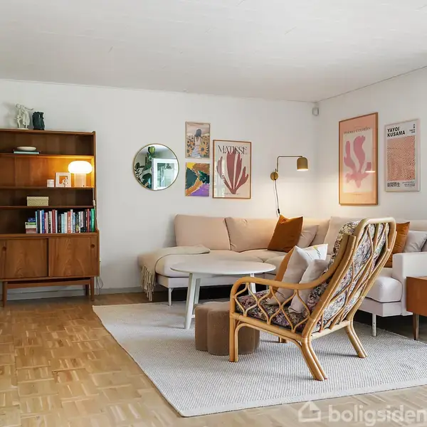 En stue med en lys beige hjørnesofa omgivet af diverse vægkunst. En træreol med bøger står ved siden af. En lænestol og grøn plante fuldender indretningen.
