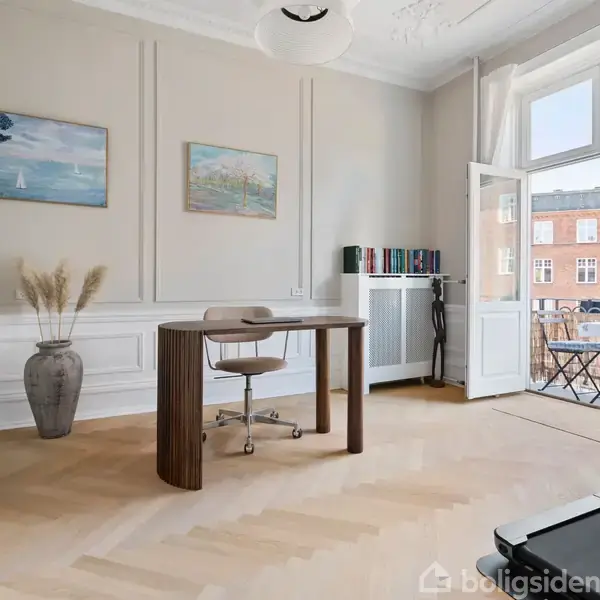 Et skrivebord med en stol står stille i et lyst rum med trægulv. To malerier hænger på væggen, og der er adgang til en balkon med udsigt over bygninger.