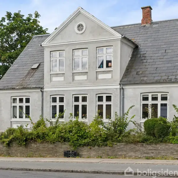 Hvidt, to-etagers hus med kviste og skråtag står langs en vej, omkranset af en lav stenmur og grønne buske. Klar himmel og træer i baggrunden.