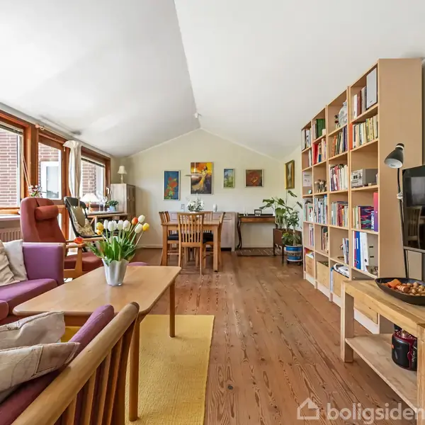 En stue med sofa og bord, dekoreret med blomster. Reol fyldt med bøger, og et tv hænger på væggen. Sollys strømmer ind gennem store vinduer.