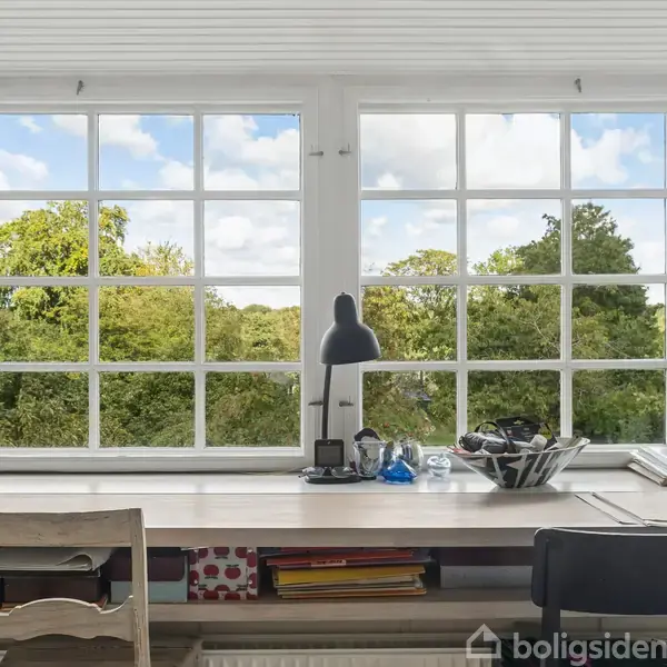 Et skrivebord står foran store vinduer med udsigt til træer og himmel. På bordet er der en lampe, bøger og diverse genstande. En stol er placeret ved bordet.