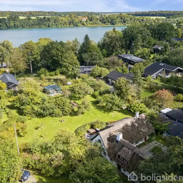Stråtækt hus omgivet af frodige træer og buske ligger i et grønt område. Flere huse ses spredt omkring, med en stor sø i baggrunden.