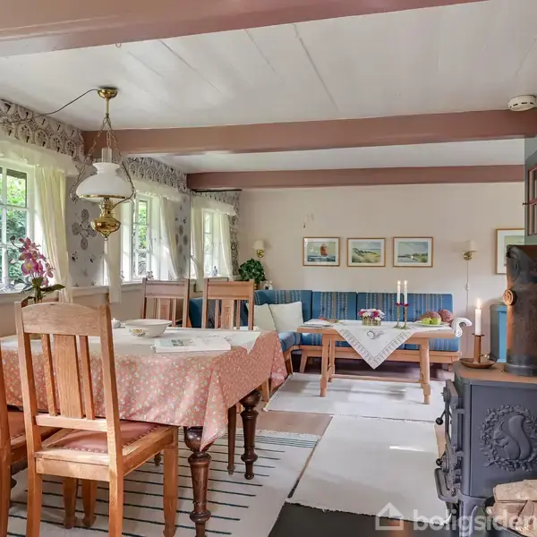 Et spisebord med fire træstole, dækket med en lærredsdug og blomster, står ved siden af en brændeovn. I baggrunden ses en sofa og flere kunstværker på væggen.