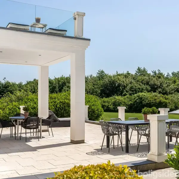 En udendørs terrasse har borde og stole arrangeret under en overdækning. Omkring terrassen er frodige grønne buske, der skaber en rolig, naturpræget atmosfære.
