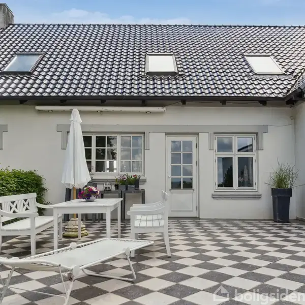 Et hvidt hus med skråtag har en terrasse med ternet flisebelægning. En hvid solseng, bord og stole står under en parasol, omgivet af grønne planter.