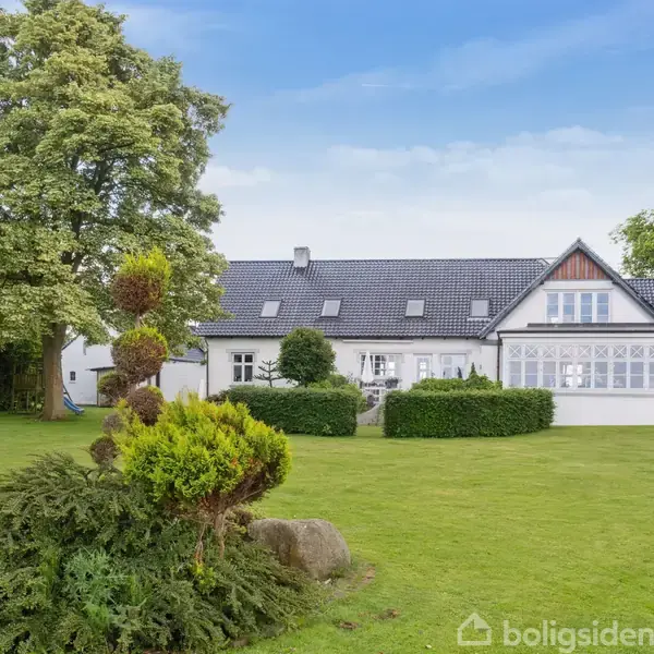 Et hvidt hus med mørkt tag står i en velplejet have med trimmede buske og store træer, omgivet af en grøn græsplæne og klar blå himmel.