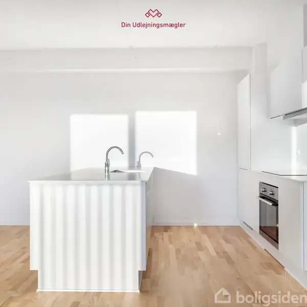 Køkkenbord med to vandhaner står i et lyst rum med trægulve. Der er en indbygget ovn og skabe i moderne, minimalistisk design. Tekst: "Din Udlejningsmægler".