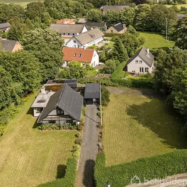 Græsplæne strækker sig gennem en have, omkranset af buske. I baggrunden ses et hus med mørkt tag på højre side og forskellige træer.