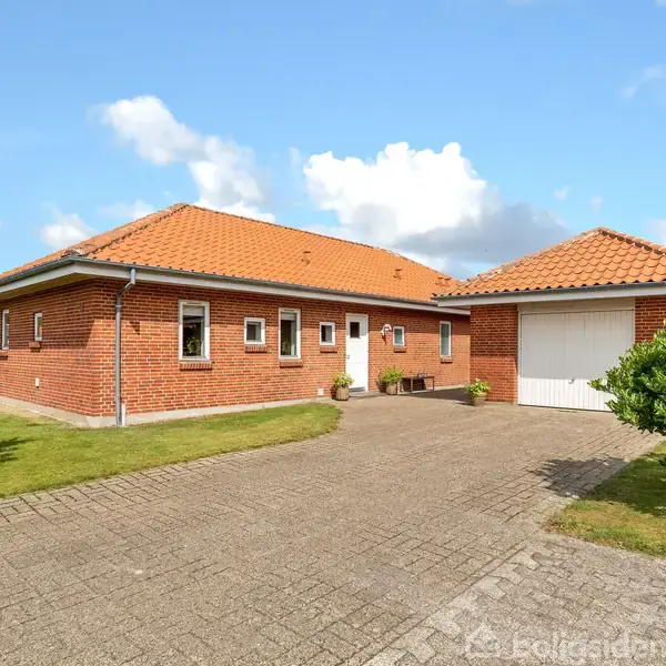 Rødt murstenshus med rødt tegltag står på en brolagt indkørsel. Ved siden af huset er der en hvid garage. Græs og buske omgiver bygningen under en blå himmel.