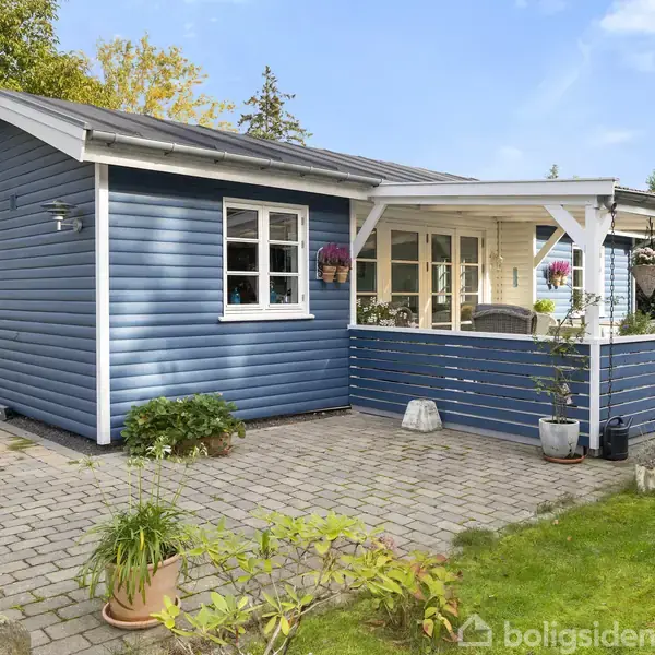 En blå træhytte med hvide vinduesrammer har en overdækket terrasse. Terrasseområdet er dekoreret med potteplanter. Omgivet af en stenbelagt gårdhave og frodig have.