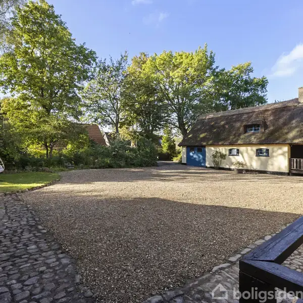 En gårdsplads med grusbelægning omgivet af to huse med stråtag; træer omkranser området, og en hængestol står til venstre. Atmosfæren er landlig og fredelig.