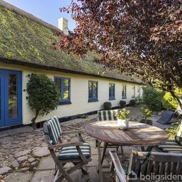 Et rundt havebord med fire stole står på en stenterrasse. Bordet står under et træ foran et langstrakt gult bindingsværkshus med stråtag og blå døre og vinduer.