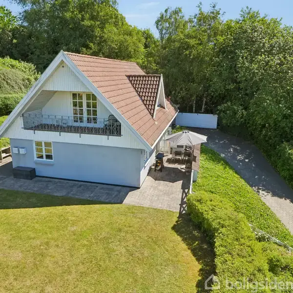Et hvidt hus med rødt tag står på en velplejet græsplæne. Foran huset er der en terrasse med et bord og parasol. Omgivet af træer og buske.