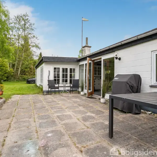 Et hus med åbne terrassedøre står ved en flisebelagt terrasse. Et bord og grill er synlige. Omgivet af en græsplæne og træer under en klar himmel.