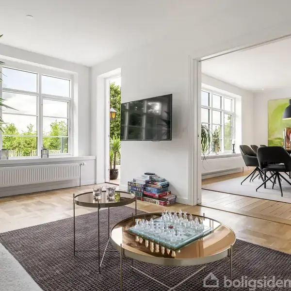 Stue med en sofa, et sofabord med skakbræt, og en tv på væggen, der åbner op til en spisestue med spisebord og stole. Store vinduer giver lys.