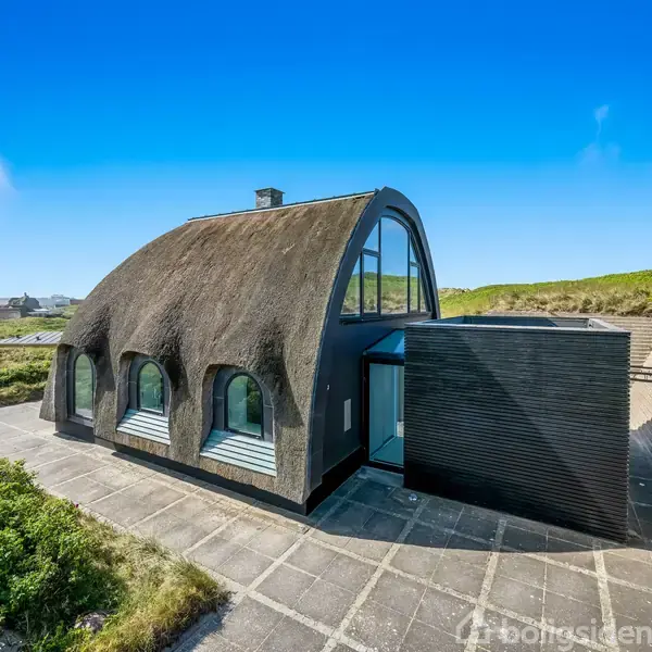 Hus med stråtag og buede vinduer står på en flisebelagt terrasse omgivet af grønt landskab under en klar blå himmel.