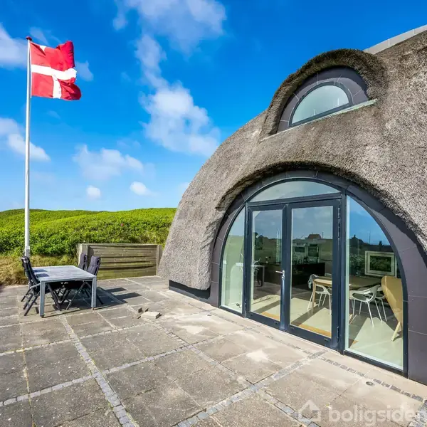 En kuppelformet bygning med store glasdøre står på en flisebelagt terrasse. Et dansk flag vajer i vinden. Bygningen ligger i et grønt landskabsområde under en blå himmel.