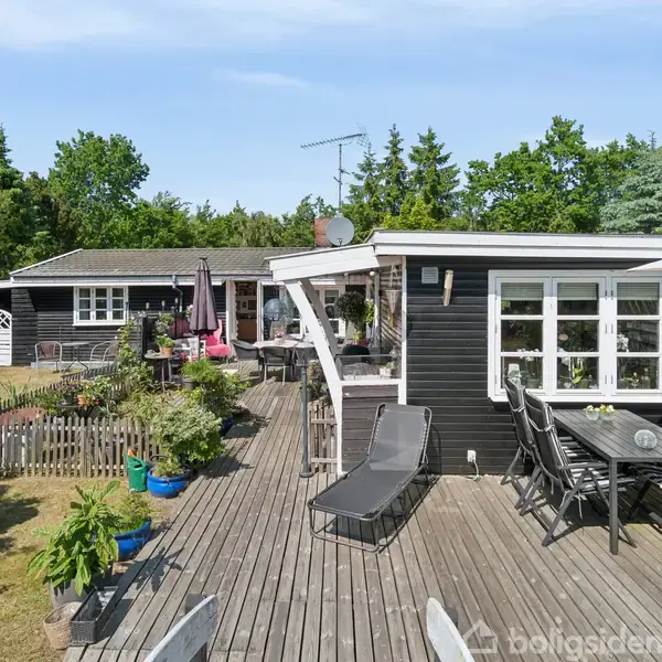 Sort sommerhus med terrasse; solstol og spisebord i forgrunden. Flere planter i krukker omkring, omkranset af græsplæne og buske. Træer i baggrunden under blå himmel.