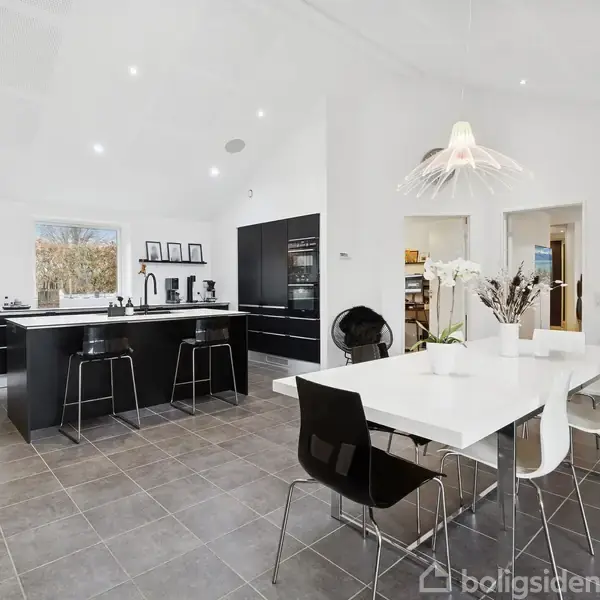 Køkken med moderne sorte skabe og en central ø omgivet af barstole. Et hvidt spisebord står i forgrunden. Rummet er lyst med store fliser og en loftlampe.