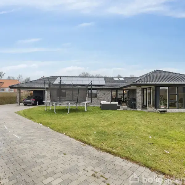 En grå murstensvilla med fladt tag står stille i en velplejet have med græsplæne. En trampolin og havemøbler er placeret på græsset. En brolagt sti fører til huset.