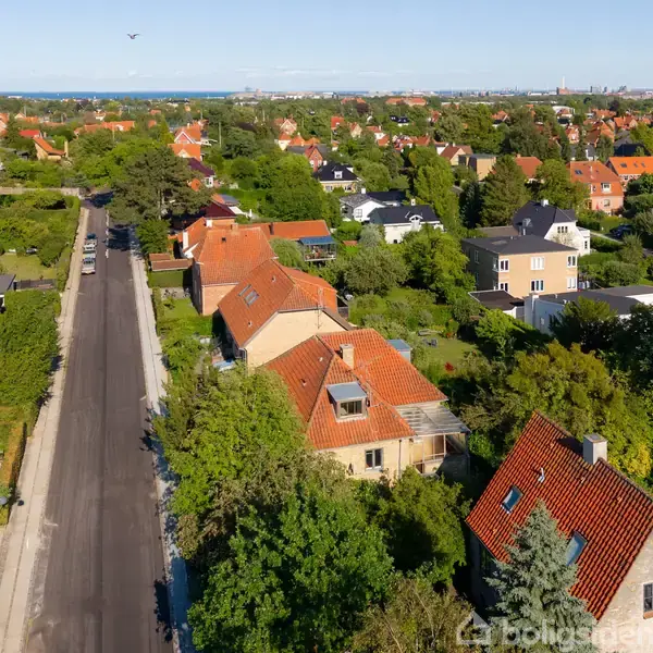 Røde hustage i en række langs en stille vej, omgivet af frodige grønne haver og træer, med flere huse synlige i det fjerne under en klar blå himmel.