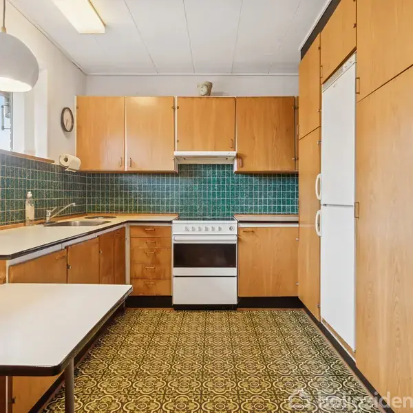 En køkken med træskabe og blå fliser omkranser en kogeplade med emhætte. En lys bordplade og et mønstret gulv tilføjer detaljer. En stor pendellampe hænger over bordet.