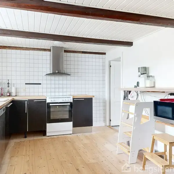 Køkken med komfur og emhætte i midten, omgivet af sorte skabe. I højre side er der en mikrobølgeovn på en køkkenbordplade, med skammel og stige i lyst træ foran. Væggene er hvide med træbjælker i loftet.