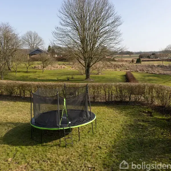 En trampolin står stille i en stor, grøn have omgivet af hække og træer. En bred plæne strækker sig bagved med flere træer og åbent landskab i baggrunden.