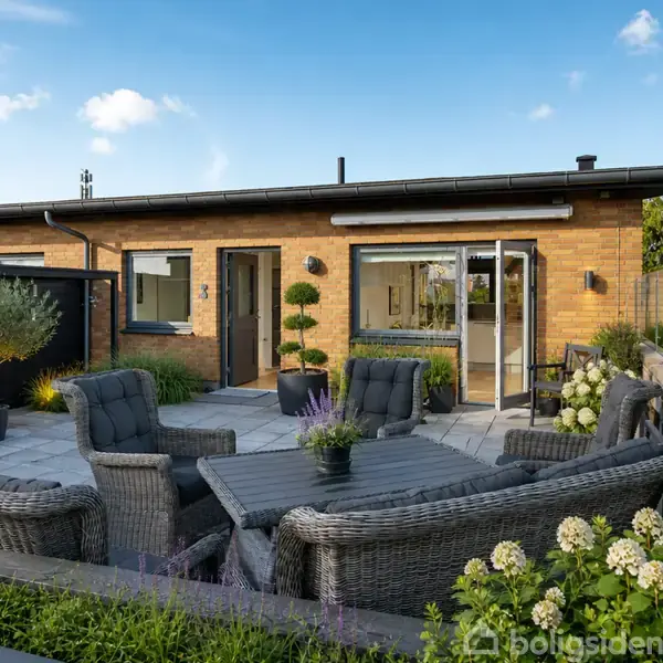 Udendørs terrasse med grå kurvemøbler og lavendel, foran en moderne murstensbygning. Solrig haveomgivelse med planter og klare blå himmel.