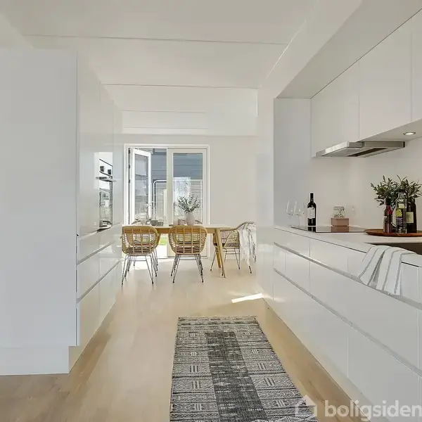Køkkenet viser en hvid indretning med moderne skabe og en bordplade. En løber ligger på gulvet. Spisestuen i baggrunden har et bord og stole, og der er dagslys fra vinduerne.