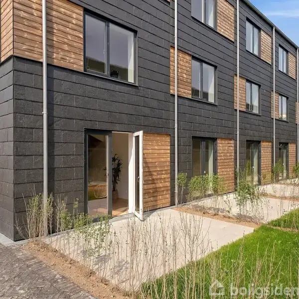 Bygning med moderne rækkehuse, åbne terrassedøre, og sort-brune facadeelementer. Beliggende i et grønt område med stier og planter.