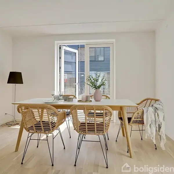 Fem spisebordstole i flet står omkring et lyst spisebord med vaser og drikkeglas. En åben dør fører til en balkon. En gulvlampe står i hjørnet.