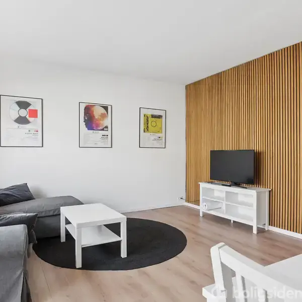 En grå sofa står i en lys stue med trægulv. Ved siden af er et hvidt sofabord på et rundt sort tæppe. Væggen har tre kunstneriske plakater. En tv-skærm er placeret på et hvidt mediemøbel foran en væg med trælister. Tre hængelamper hænger fra loftet i nærheden.